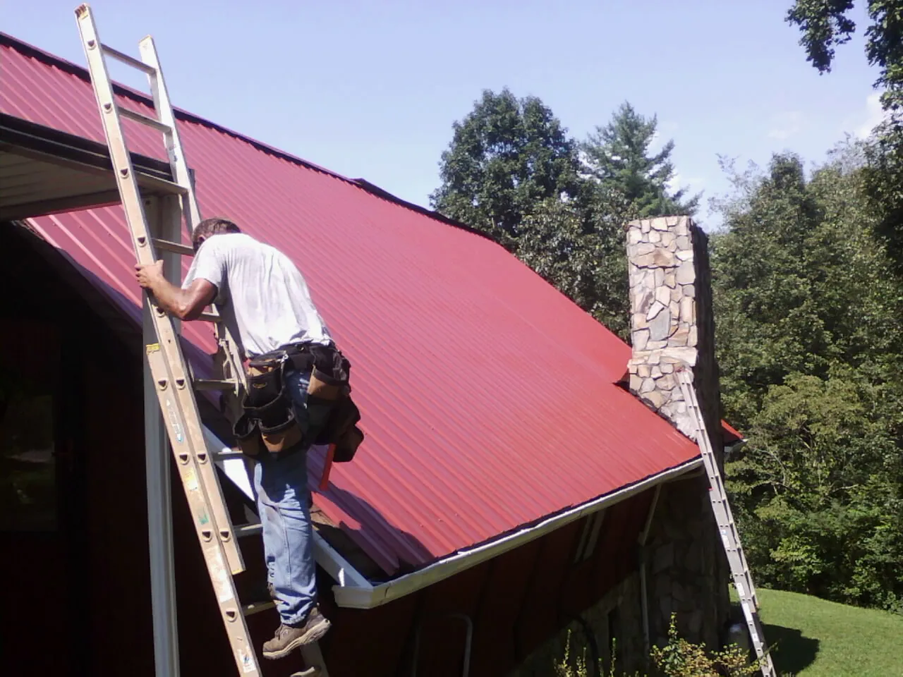 Roofer on a ladder beside a red metal roof and stone chimney — Soffit & Fascia Repair by Roofing Brothers in Norristown